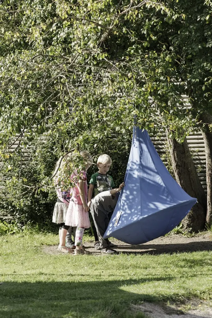 Børn og en voksen ved et blåt hængetelt på græsset en sommerdag.