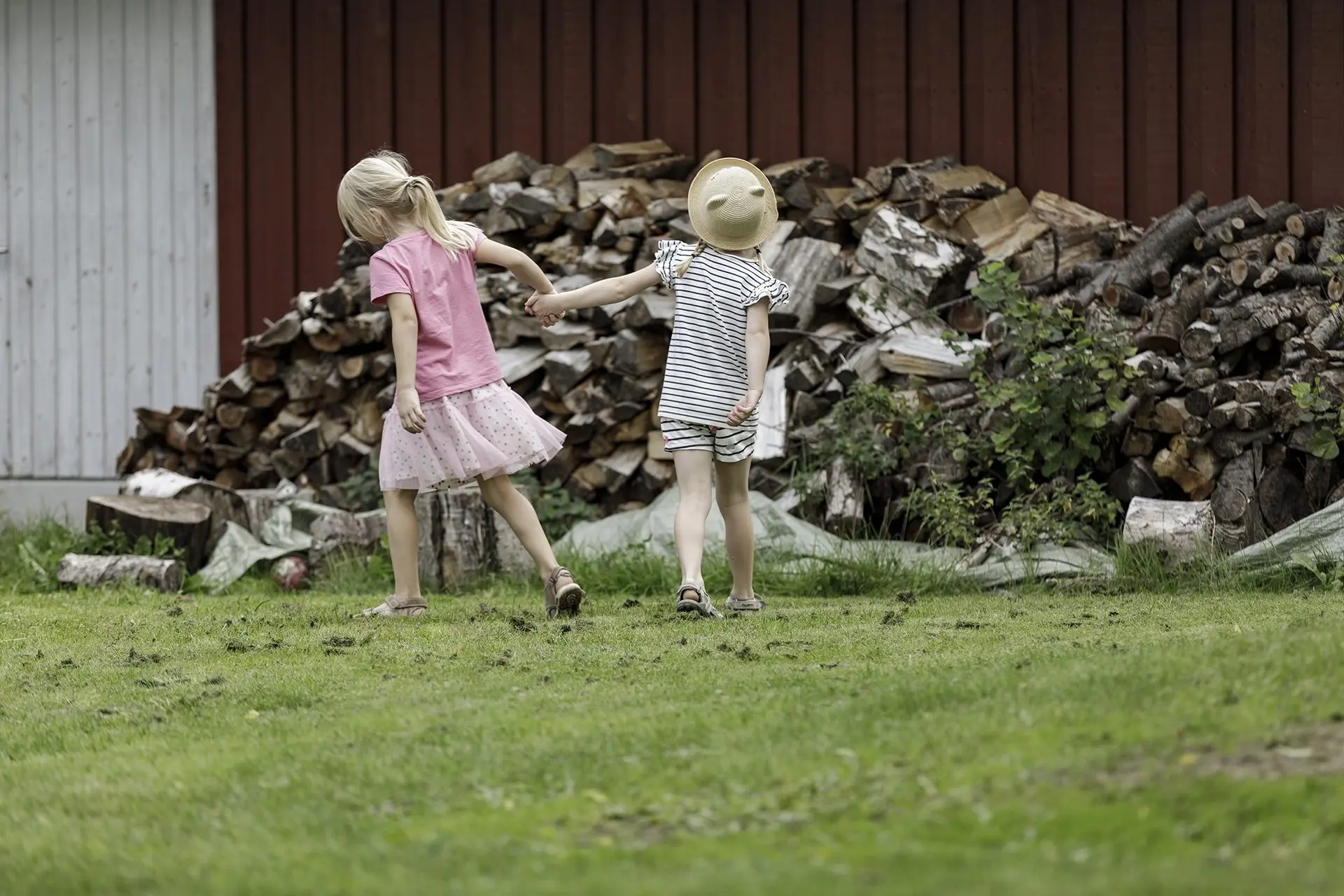 To piger går hånd i hånd over græsset forbi en stor stak brænde foran et rødt skur.