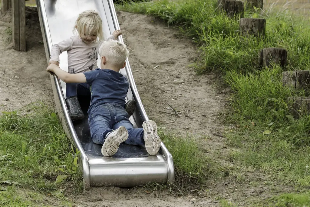 En pige i gummistøvler og en dreng i jeans og sneakers ligger på rutsjebanen i skråningen og kigger på hinanden.