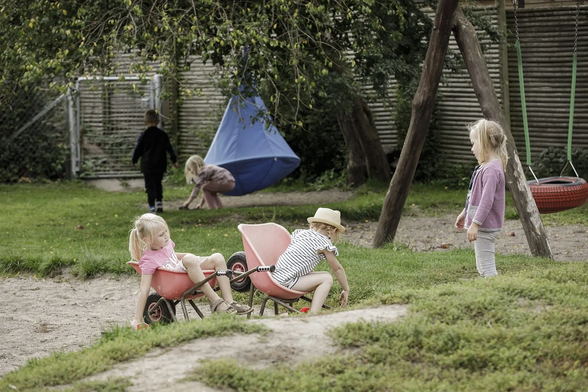 To piger sidder i hver sin lyserøde trillebør på legepladsen, mens en tredje pige ser på. Bagved hænger et blåt hængetelt og en dækgynge.